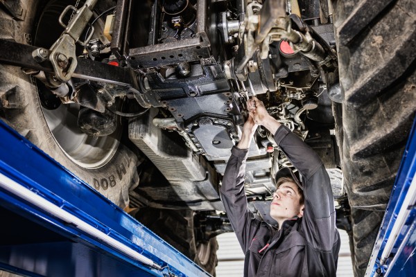 Businessfotografie zeigt Landmaschinen-Mechaniker bei der Reparatur am Unterboden eines grossen Traktors.